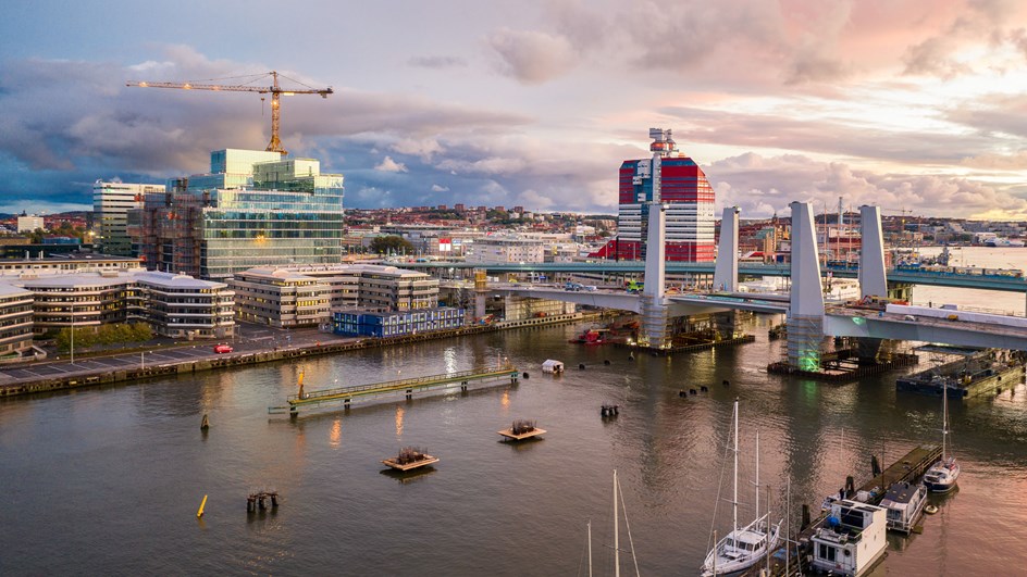 2016 fick Skanska i uppdrag av Göteborgs stad att riva den gamla uttjänta Götaälvbron från 1939 och bygga en ny bro. Hisingsbron. Bro med två pylon-par sett snett ovanifrån. I bakgrunden syns stadens siluett med byggnader och byggkran, i förgrunden vatten och båtar.