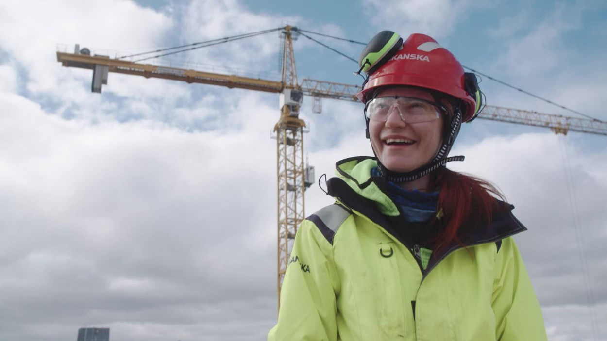 Ung kvninna i varselkläder och röd hjälm som skrattar.  I bakgrunden  avtecknar sig en byggkran mot himlen.