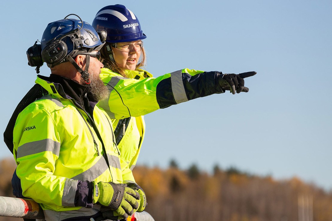 En kvinna ocoh en man i profil iklädda gula varselkäder och röda hjälmar. Kvinnan pekar framåt. 