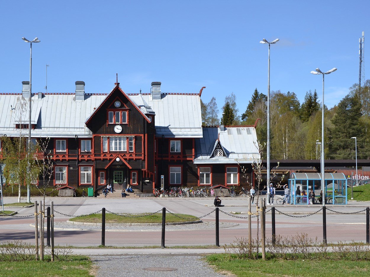 Stationshus i gammal stil med brun träfasad samt röda detaljer. Framför syns personer och en cykelparkering.