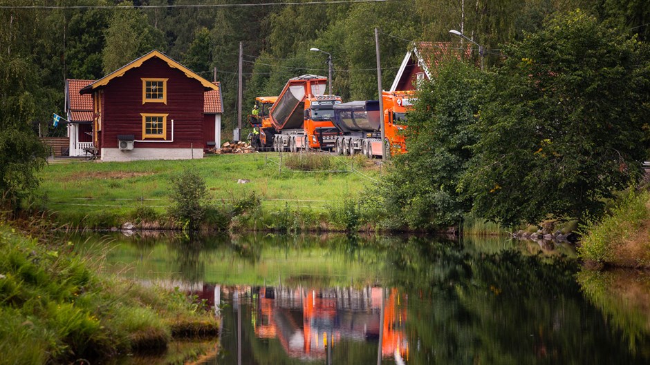 Röda stugor med vatten framför asfaltläggningsmaskiner. I bakgrundern syns tät barrskog.