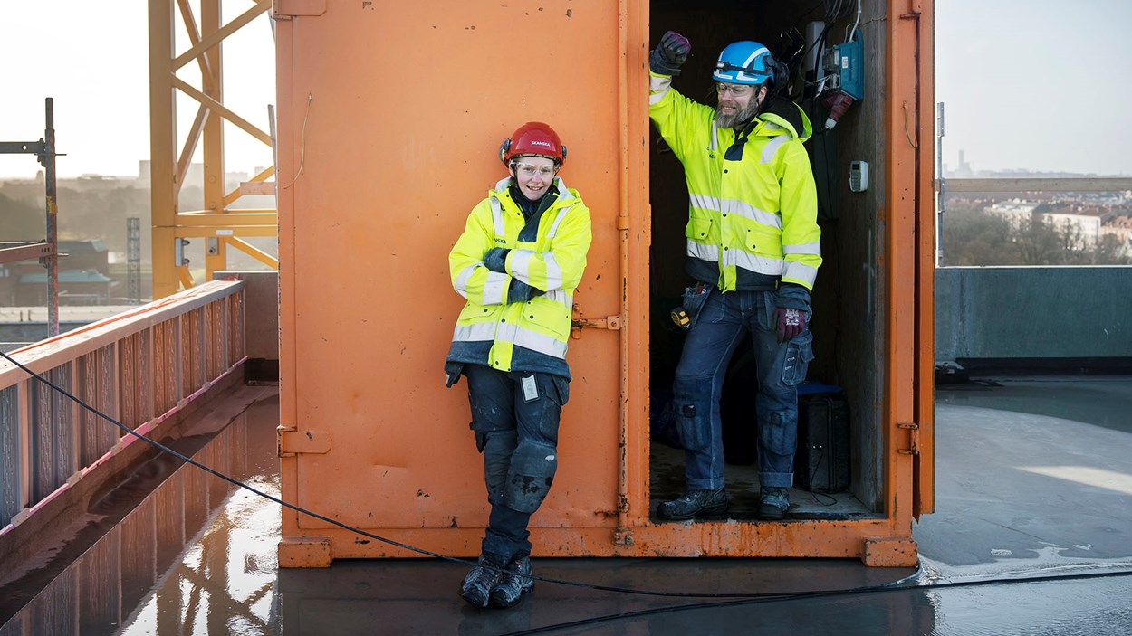 Ung kvinna och äldre man i varselkläder framför container på tak