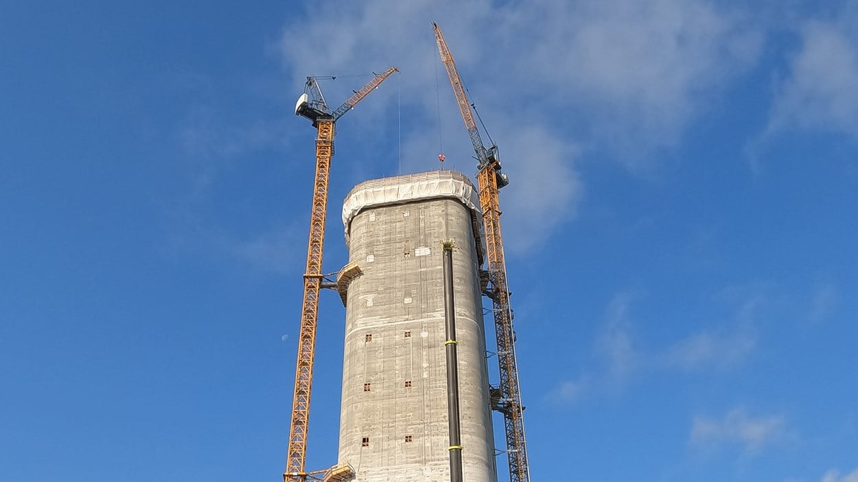 Högt torn i betong mot blå himmel.  Höga kranar på varsisn sida om tornet.