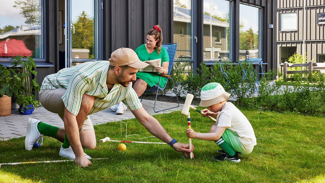 En pappa med sin lilla son spelar krocket på gräsmatten framför ett hus. I bakgruden syns mamman som sitter och läser i en trädgårdsstol.